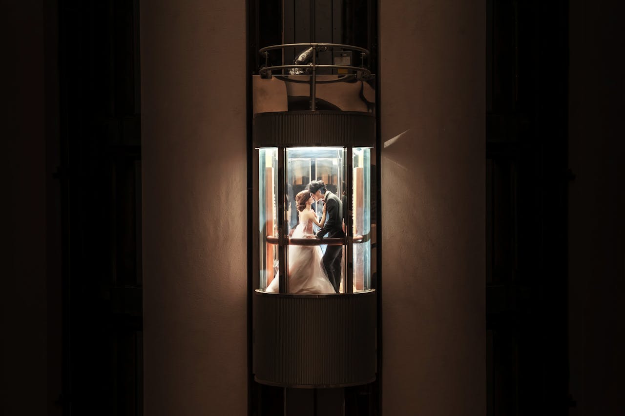 A newlywed couple shares a romantic kiss inside an illuminated glass elevator, embracing love and unity.