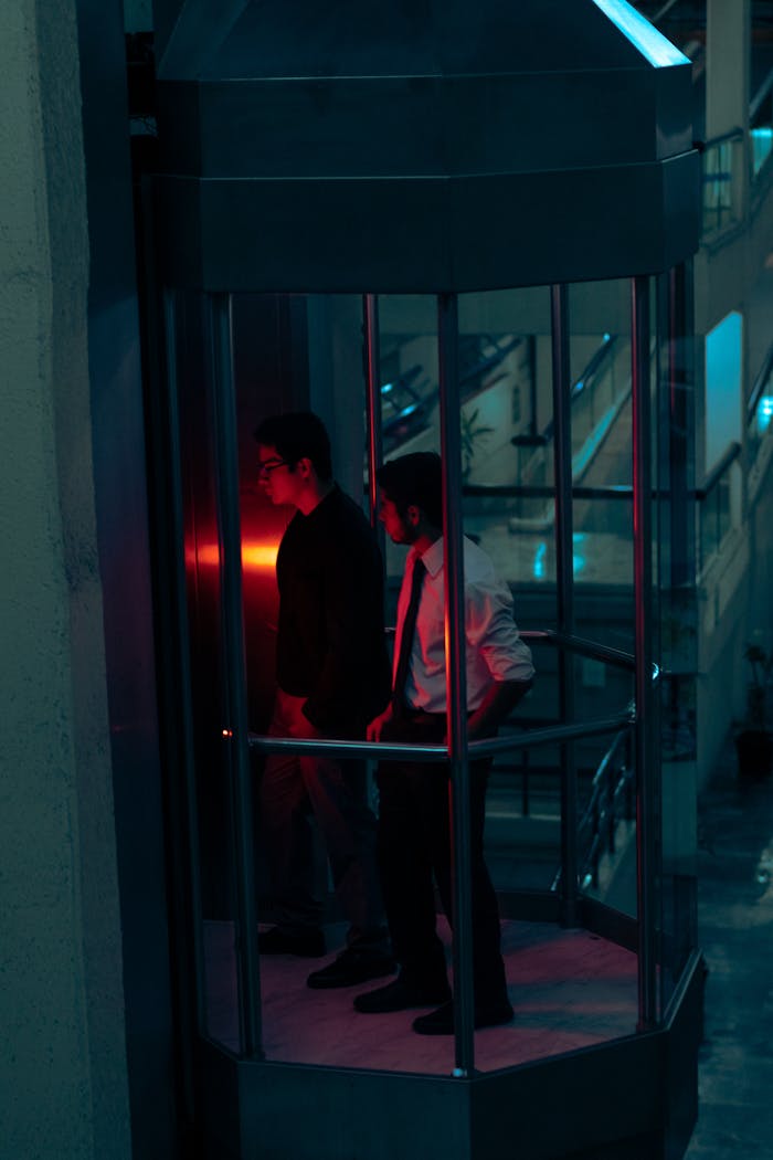 Two men in a stylish glass elevator inside a city building, captured at night.