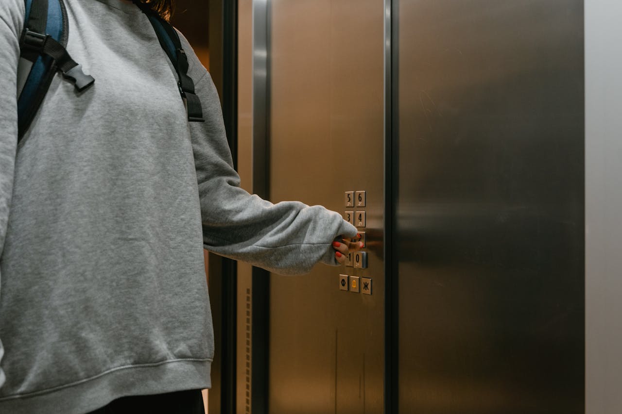 Person in grey sweater pushing elevator button with backpack in a modern building.